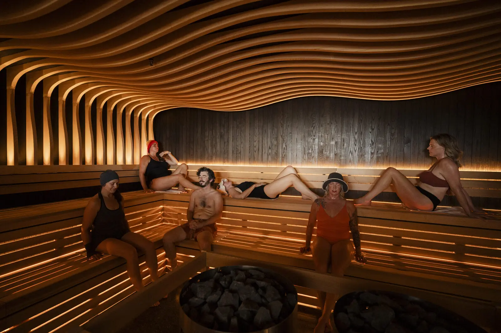 Six bathers sitting and reclining on lit tiered wooden benches under a wavy slatted wooden ceiling, with two round heaters full of stones in the foreground