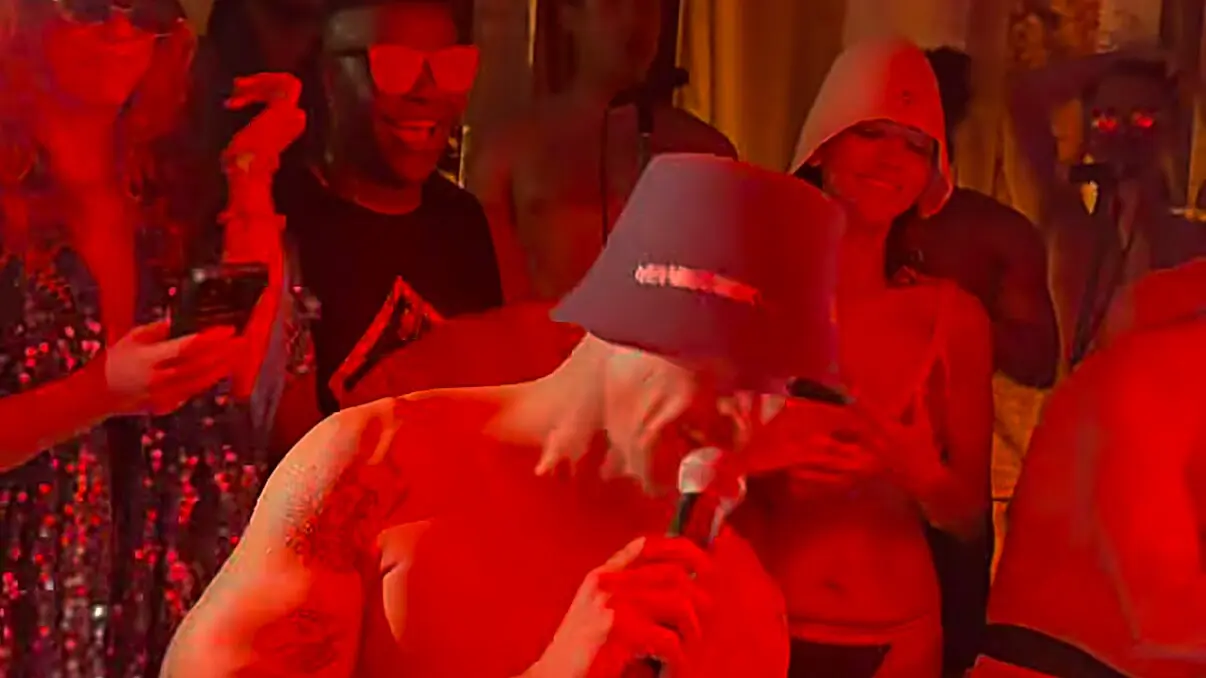 A shirtless DJ in a dark bucket hat sings into a microphone in a red-lit sauna as guests in swimwear stand behind him, some holding up phones