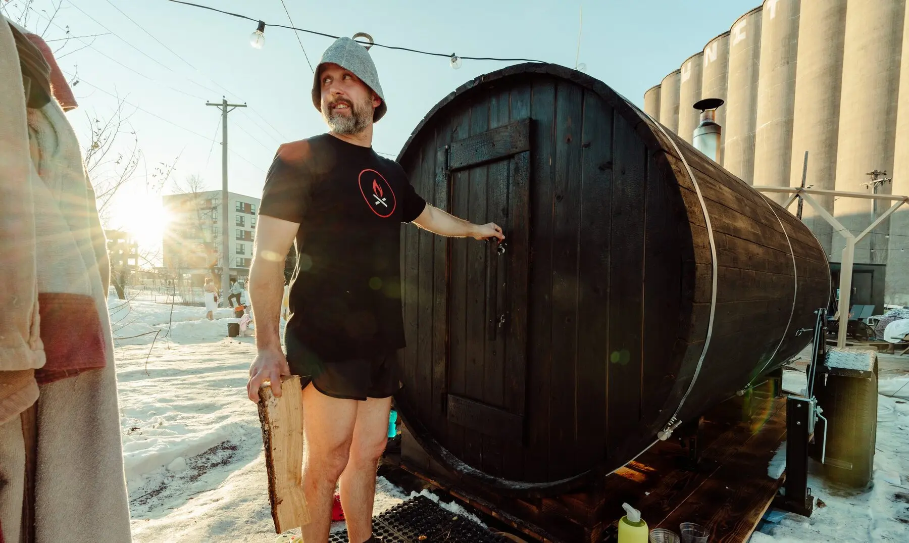 A man in a felt sauna hat holding a split log stands at the door of a dark barrel sauna on a trailer, with snow on the ground, string lights overhead and grain silos behind