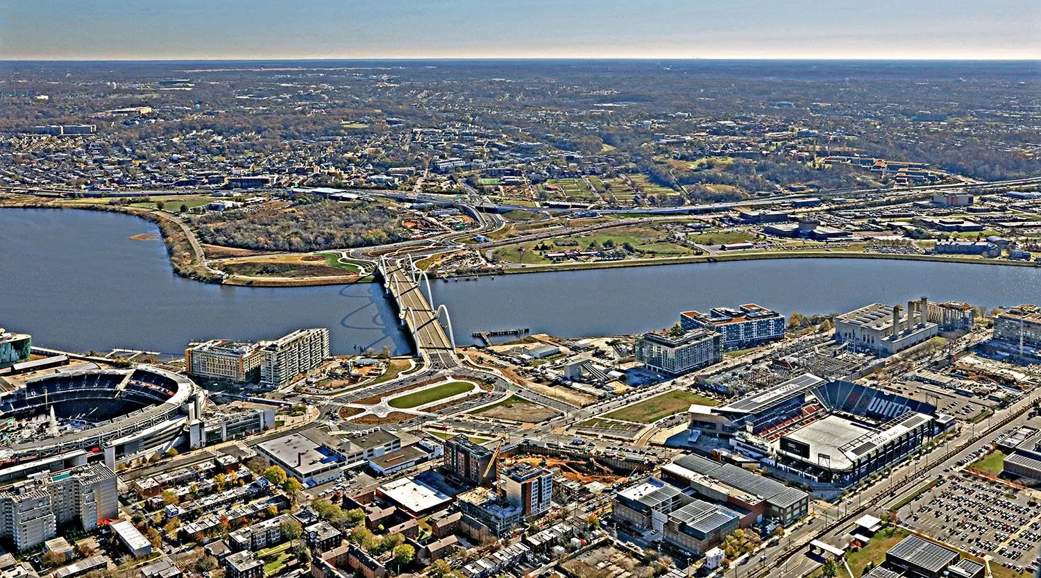 Aerial view across the Anacostia River from above Nationals Park and Audi Field to the wooded Poplar Point peninsula beside the arched Frederick Douglass Memorial Bridge