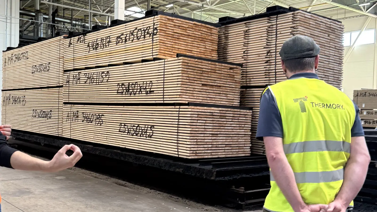 Packs of sawn boards stacked in a factory hall with batch numbers and sizes chalked on the ends, a visitor in a Thermory vest looking on