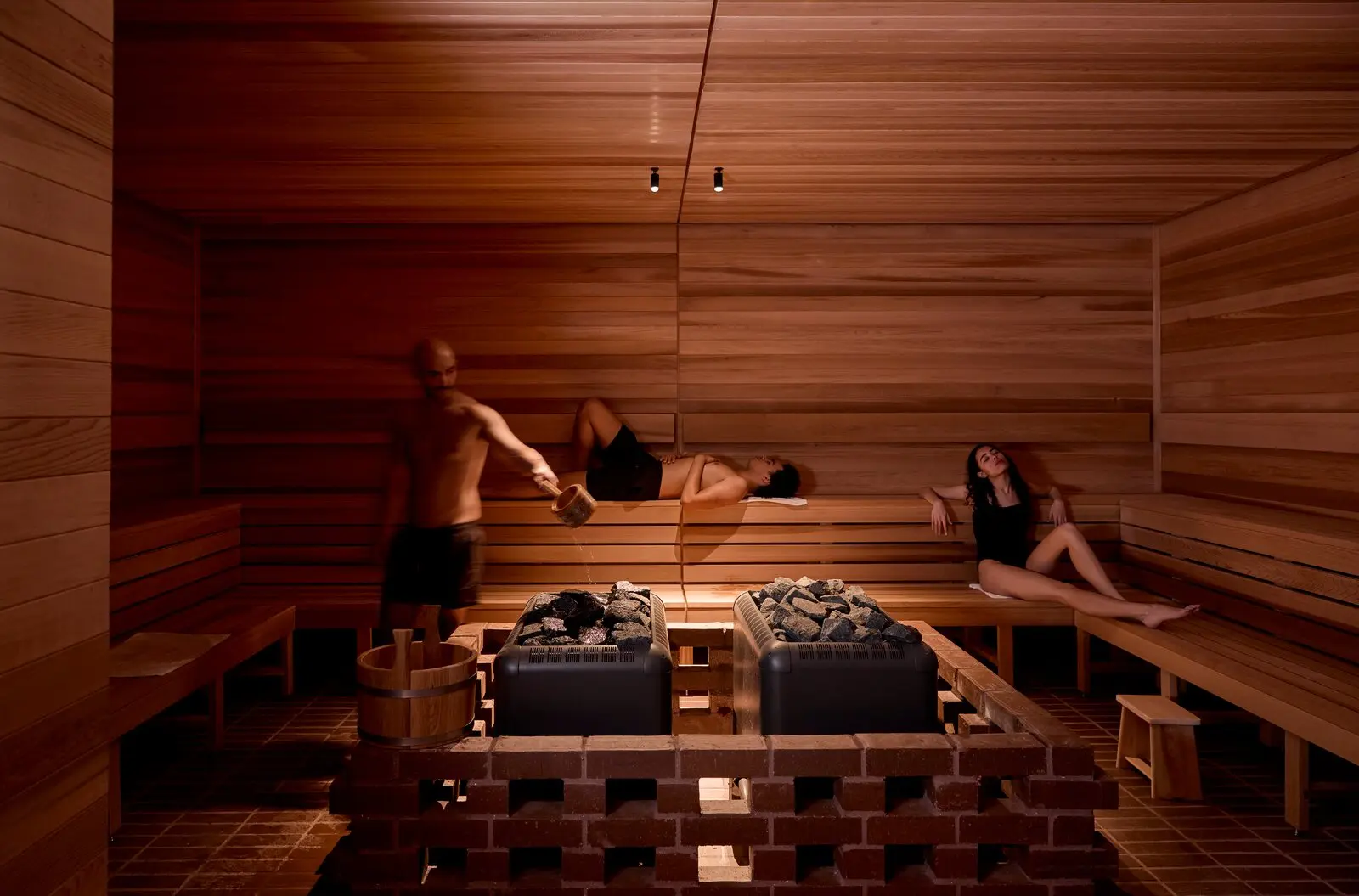 A man pours water from a wooden ladle onto one of two heaters set side by side in a low brick surround, while bathers lie and sit on the wooden benches around them