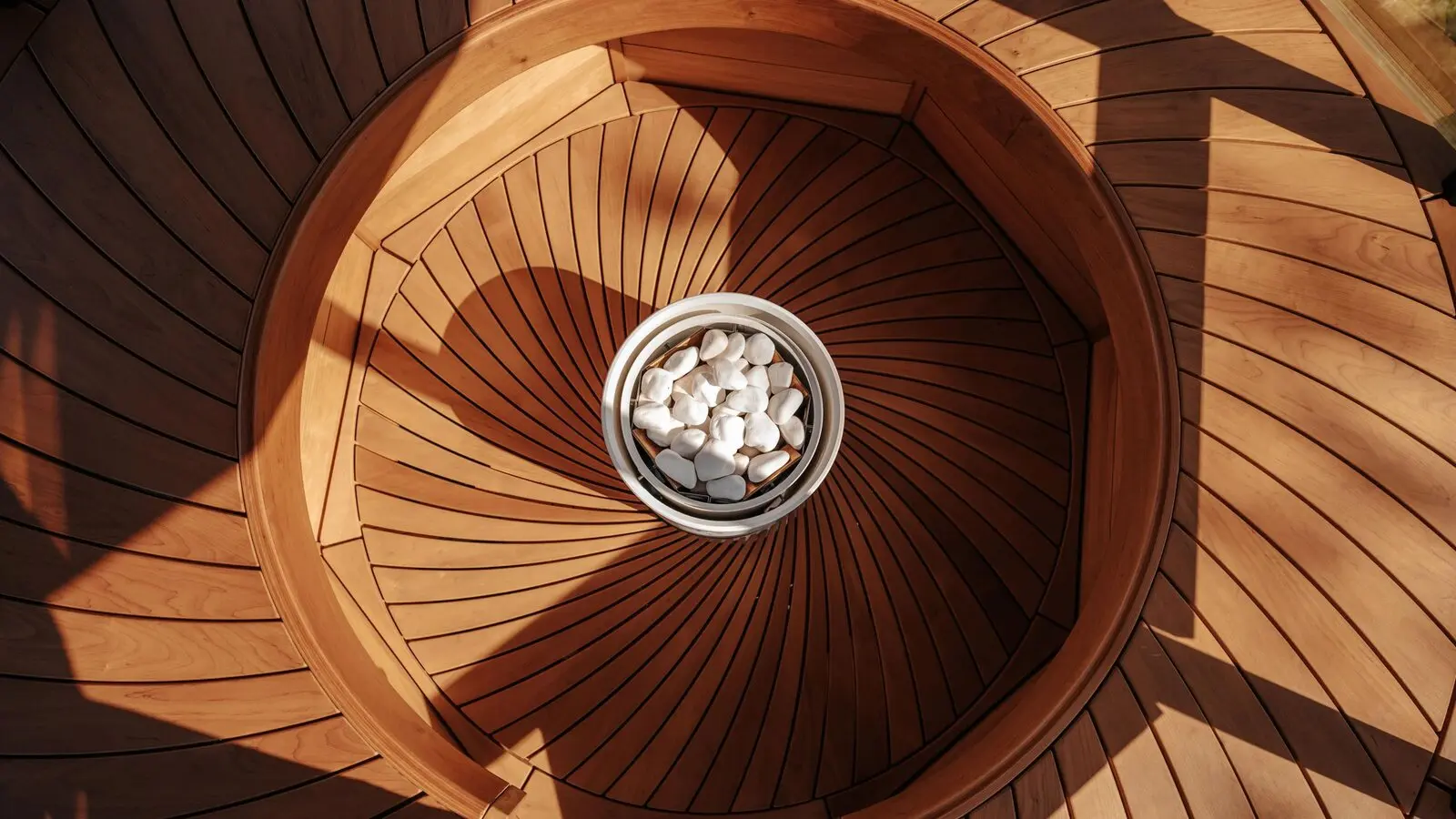 Looking down into a round wooden sauna with curved benches, a white cylindrical Tulikivi Pyry heater filled with white stones at the center