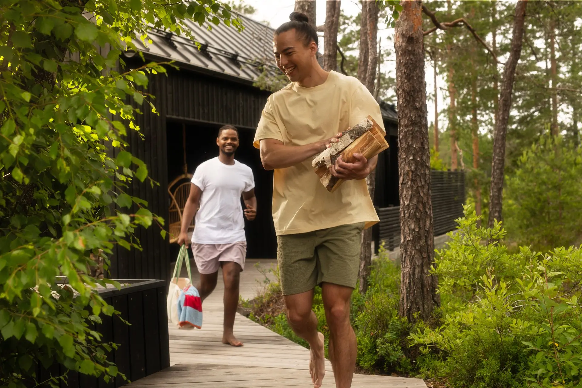 Two bathers carrying towels and firewood along a wooden path through the trees toward a dark sauna building