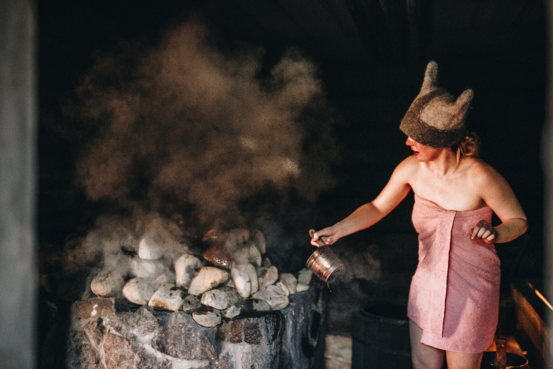 A bather ladles water onto the stones of a southern Estonian smoke sauna as steam rises