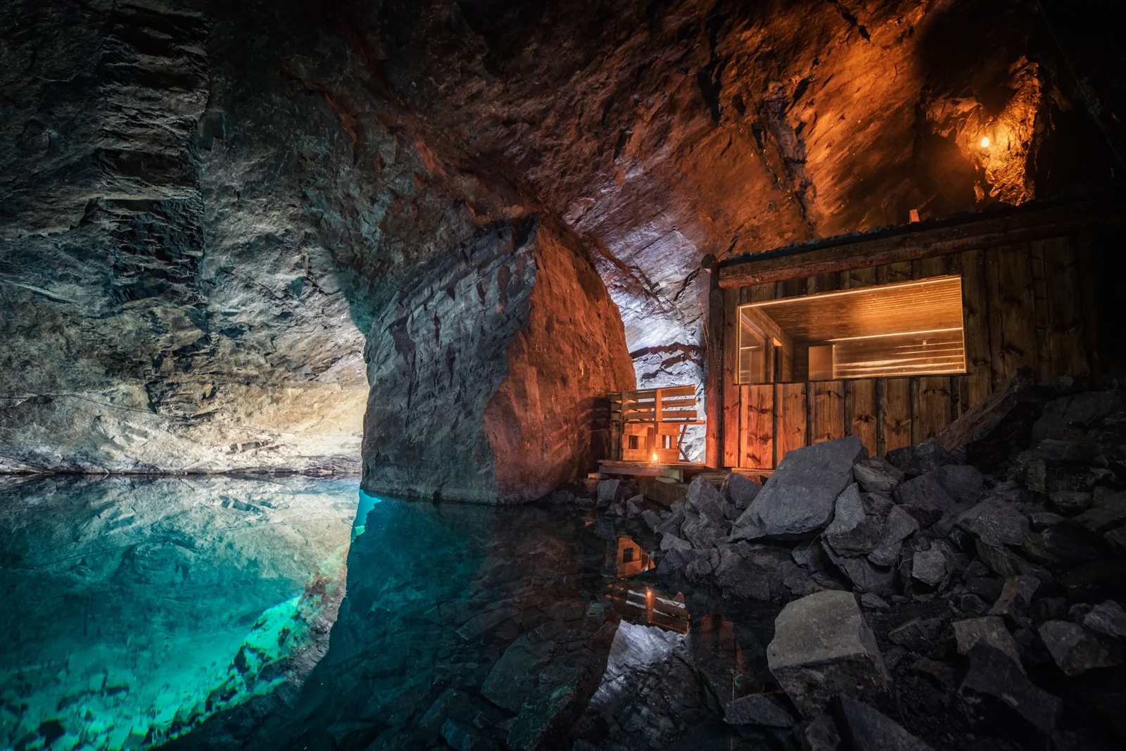 A lit wooden sauna beneath a rock wall beside turquoise underground water