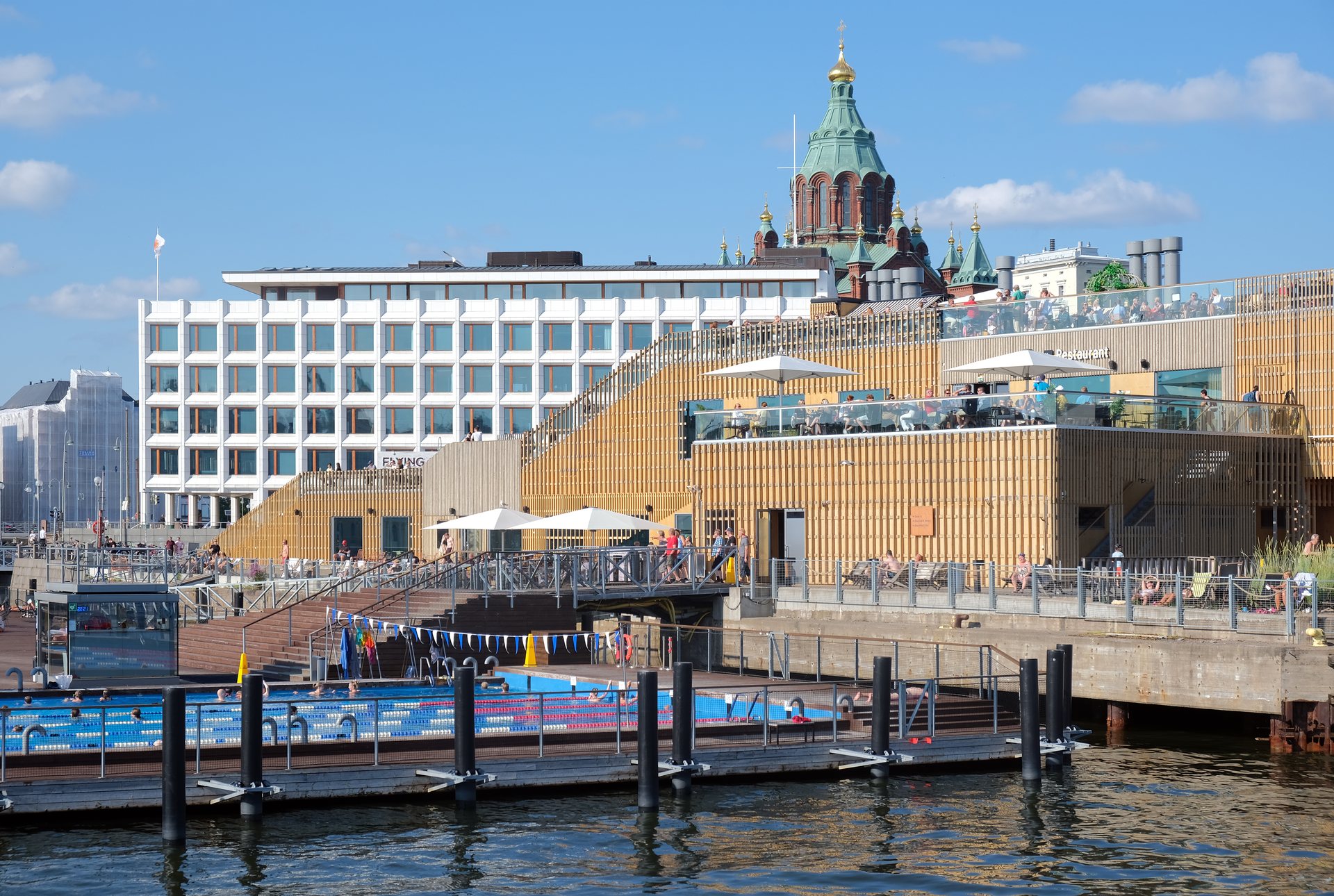 Swimmers in an outdoor pool at Allas Sea Pool on the Helsinki waterfront, with wooden buildings and a red-brick cathedral behind
