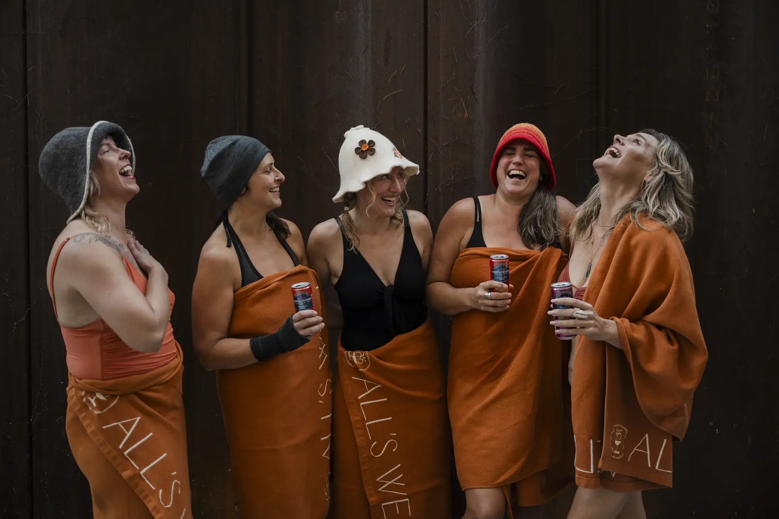 Five laughing women in swimsuits, felt sauna hats and rust-colored All's Well towels standing against a dark wooden wall, holding canned drinks