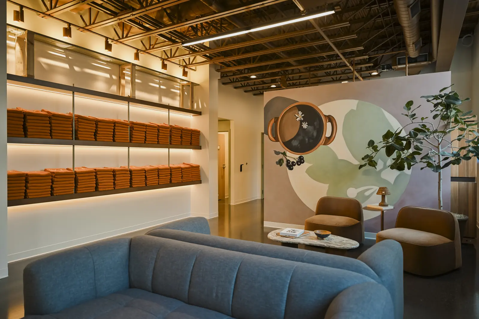 A lobby lounge with a curved blue sofa and two rust armchairs, lit shelves stacked with rust-colored towels on the left and a large painted mural on the back wall
