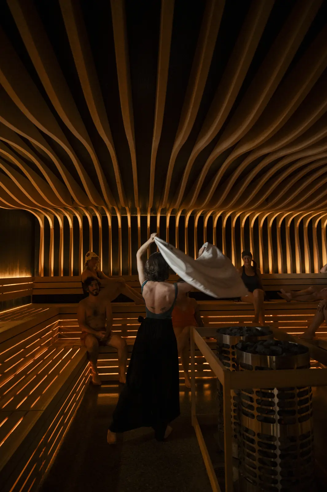 A woman in a long black dress swings a white towel overhead in a large sauna with tiered benches, a curving slatted wooden ceiling and two tall cylindrical heaters
