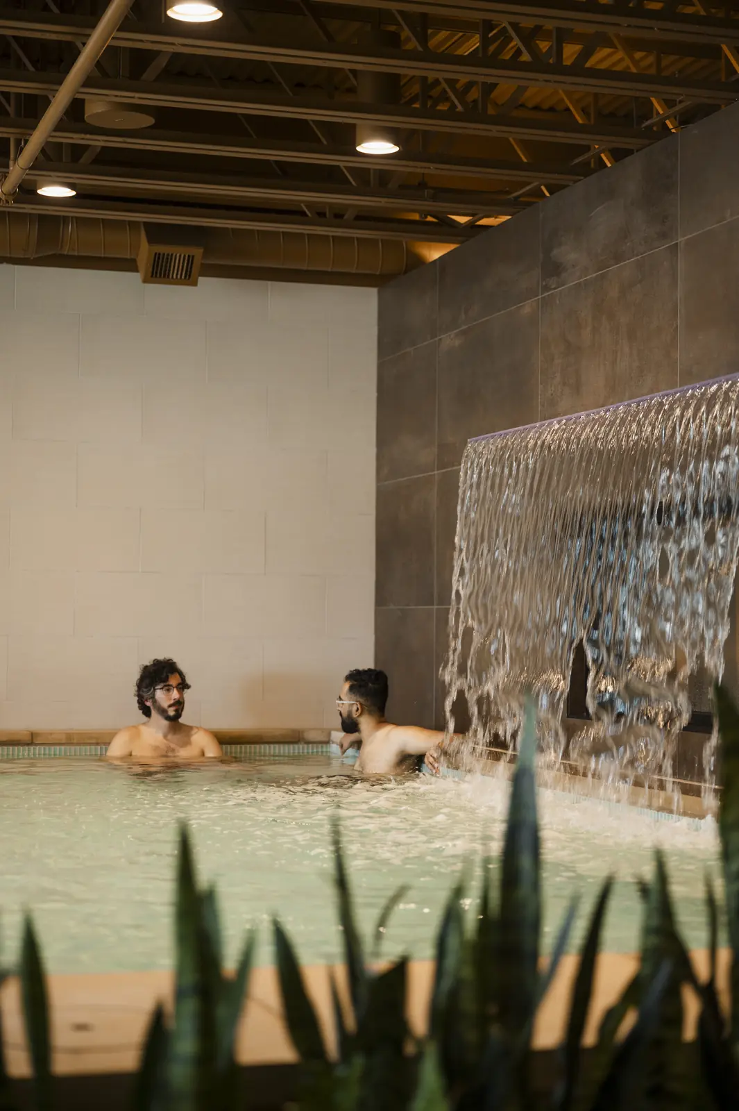 Two men talking in a pale green warm pool beside a sheet waterfall pouring from a dark tile wall, with plants in the foreground