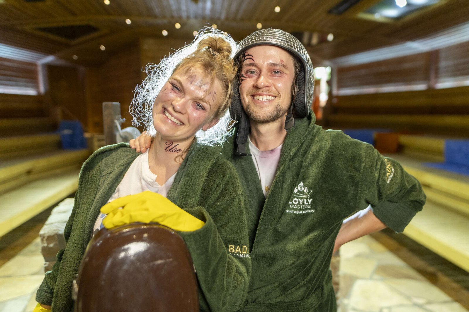 Two sauna masters in green robes and stage makeup, one wearing a mesh helmet, smile inside a large tiered sauna