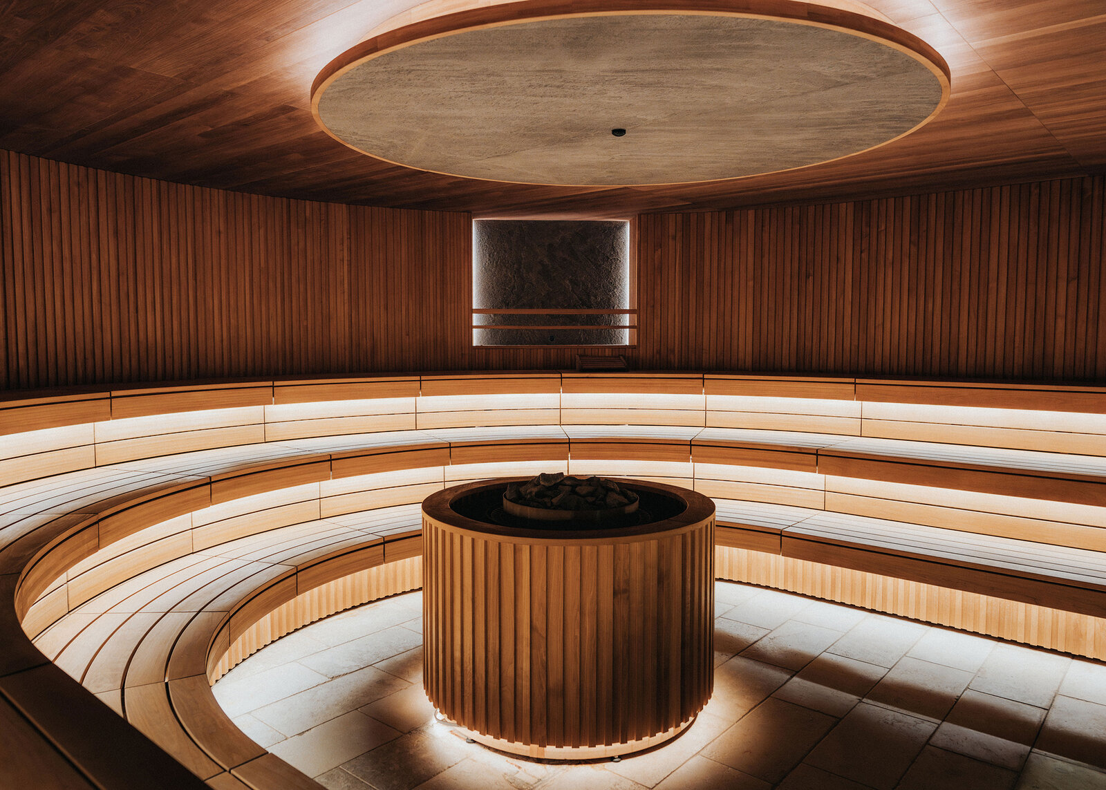 A large round commercial sauna with tiered, curved wooden benches lit from beneath, circling a tall cylindrical heater under a round ceiling panel