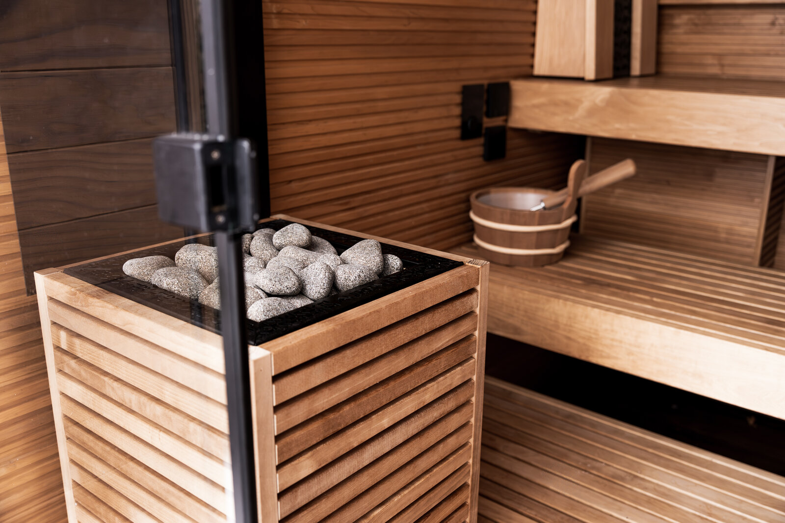 Inside an Auroom sauna, a HUUM CORE heater in a slatted wooden casing filled with stones beside wooden benches, with a wooden bucket and ladle on the bench