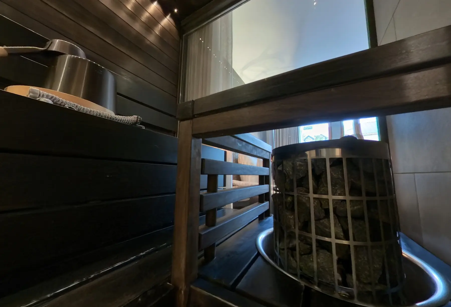 Kivillä täytetty kiuas, tummat puulauteet ja lasiseinä Lapland Hotels Bulevardin tyhjässä huonesaunassa