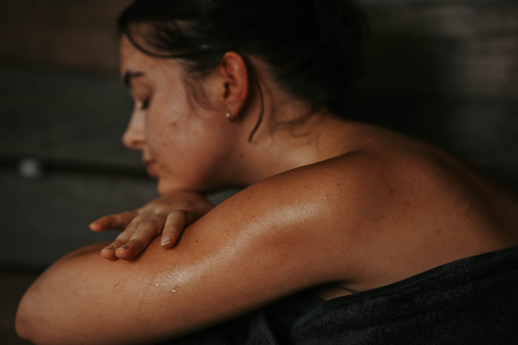 A woman wrapped in a dark towel rests her arms on a wooden sauna bench
