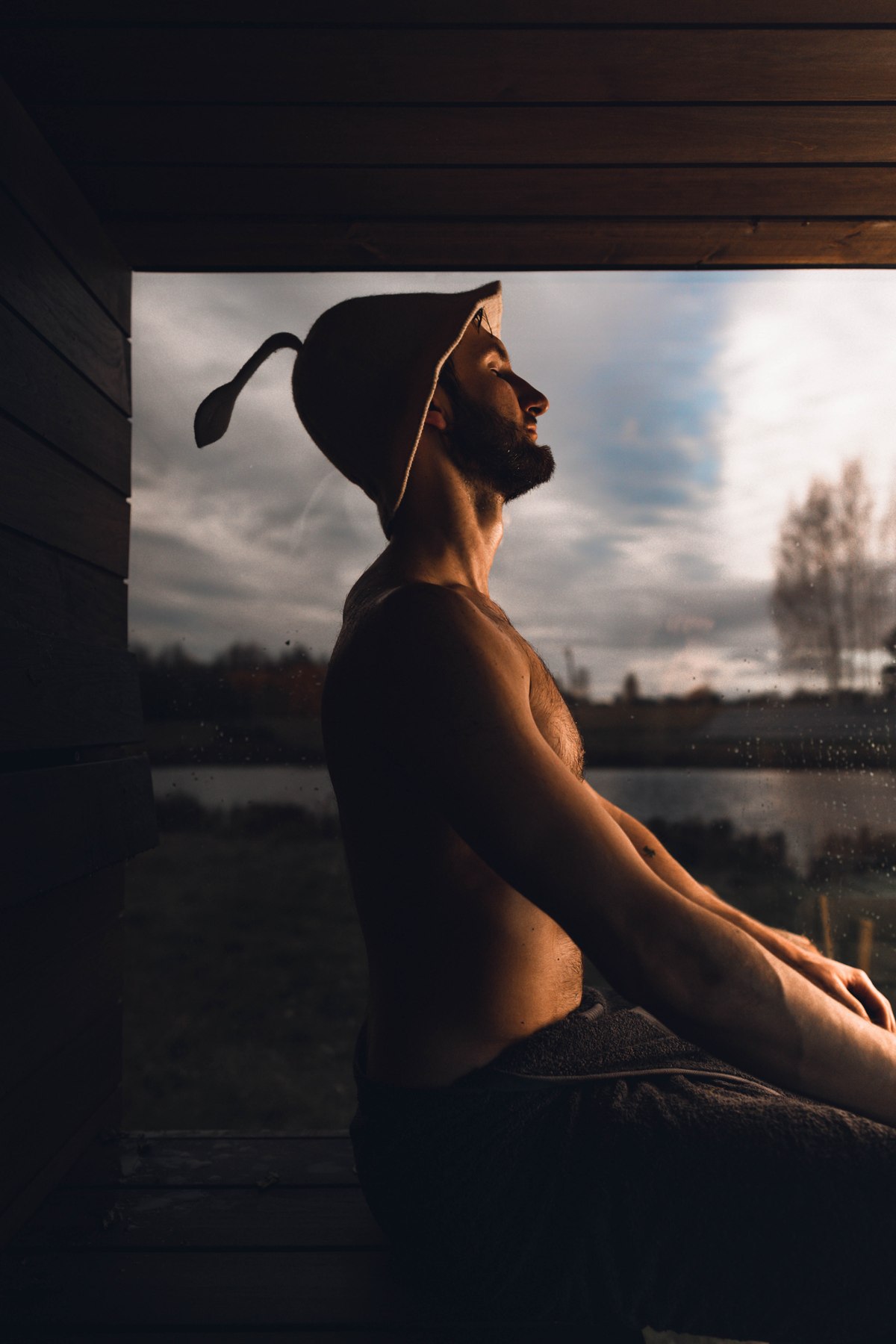 A bather wearing a sauna hat sits beside a large sauna window