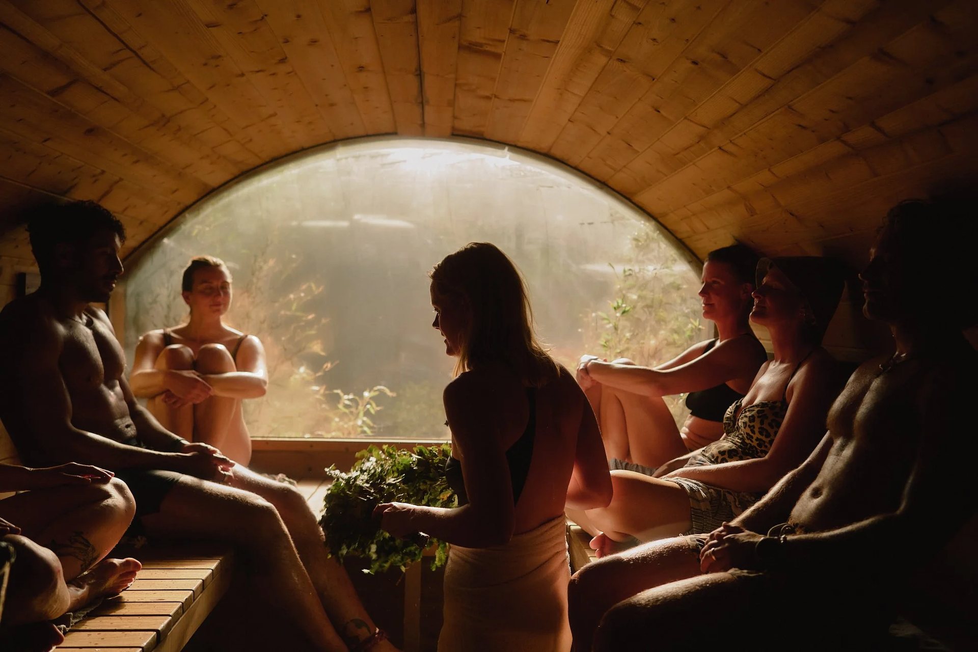 Bathers on wooden benches in a sauna with an arched window, as a woman in the center holds a leafy birch whisk