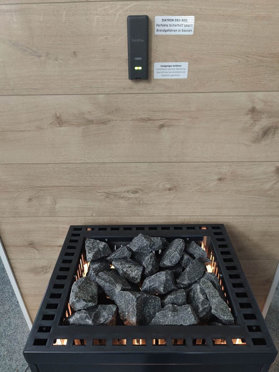 A black DIATRON OSU-400 module with a lit green status light mounted on a wood-paneled wall above a black sauna heater filled with stones