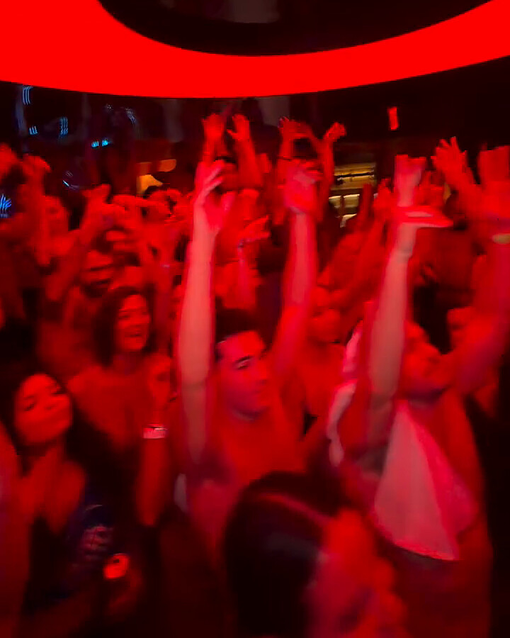 A crowd in swimwear with arms raised under red light, beneath a glowing red ring in the ceiling