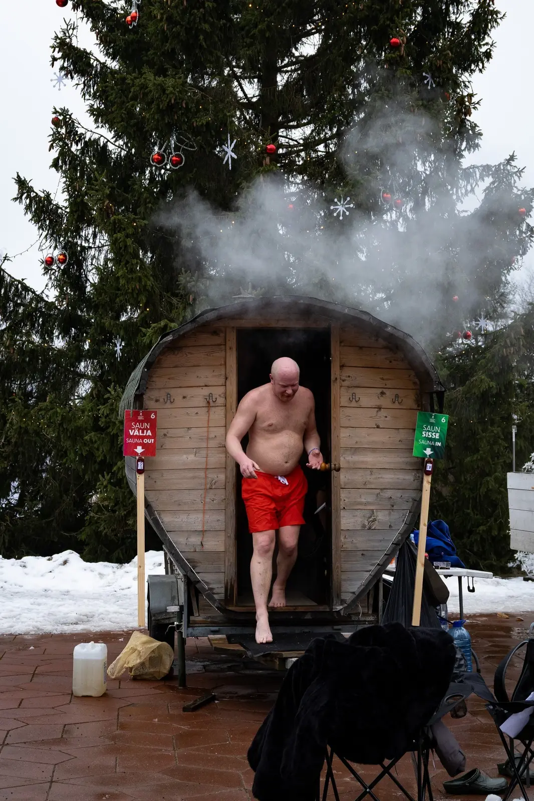Steam pouring from the roof of a barrel sauna as a man in swim trunks steps out of its door beside a snowy spruce tree