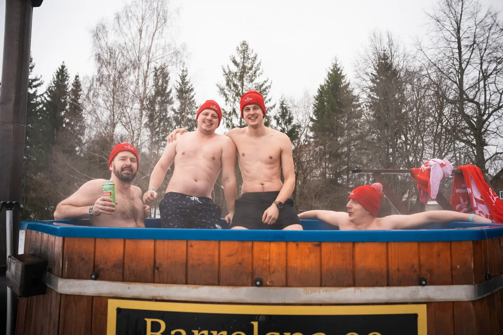 Four laughing men in red knit hats in a wood-fired hot tub, one raising a drink