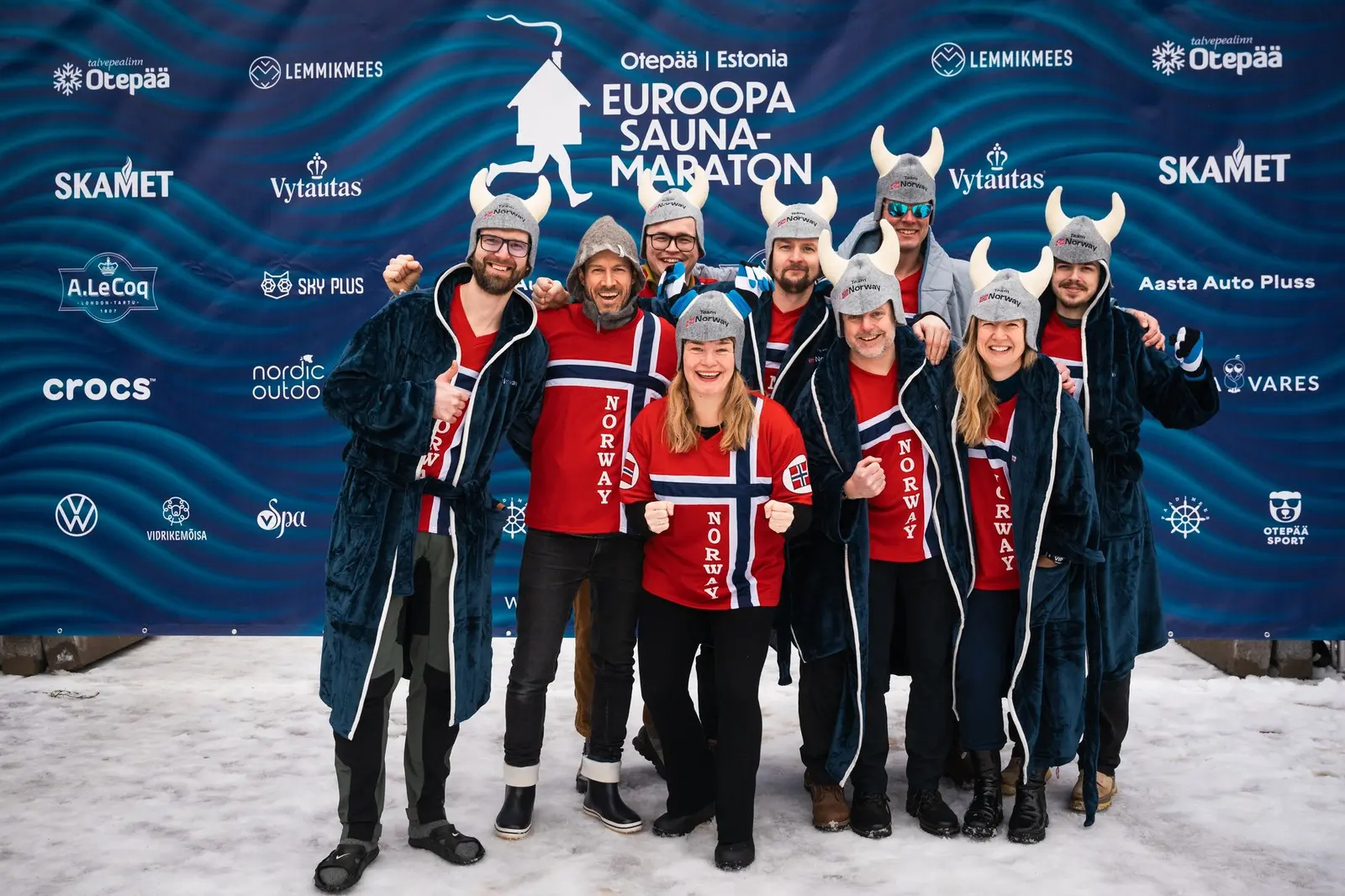 A team in red robes and capes printed with the Norwegian flag and horned Viking helmets posing in front of a European Sauna Marathon sponsor wall in the snow