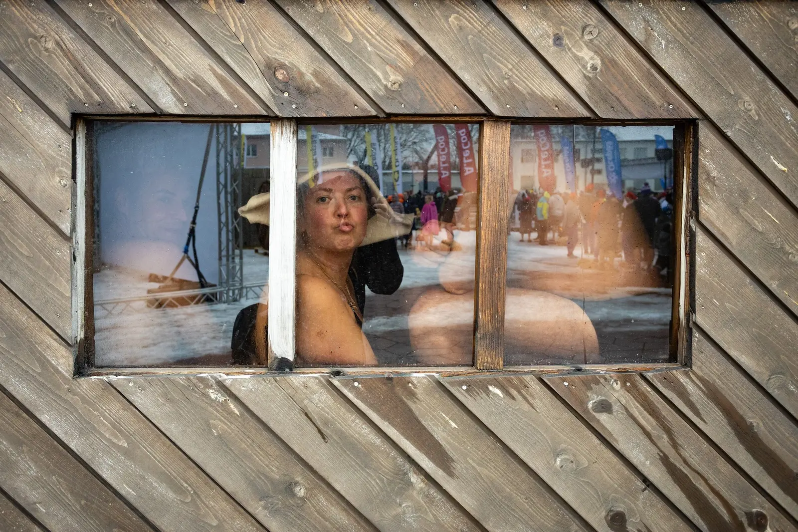 A woman in a sauna hat seen through the small window of a weathered wooden sauna, with other racers reflected in the glass