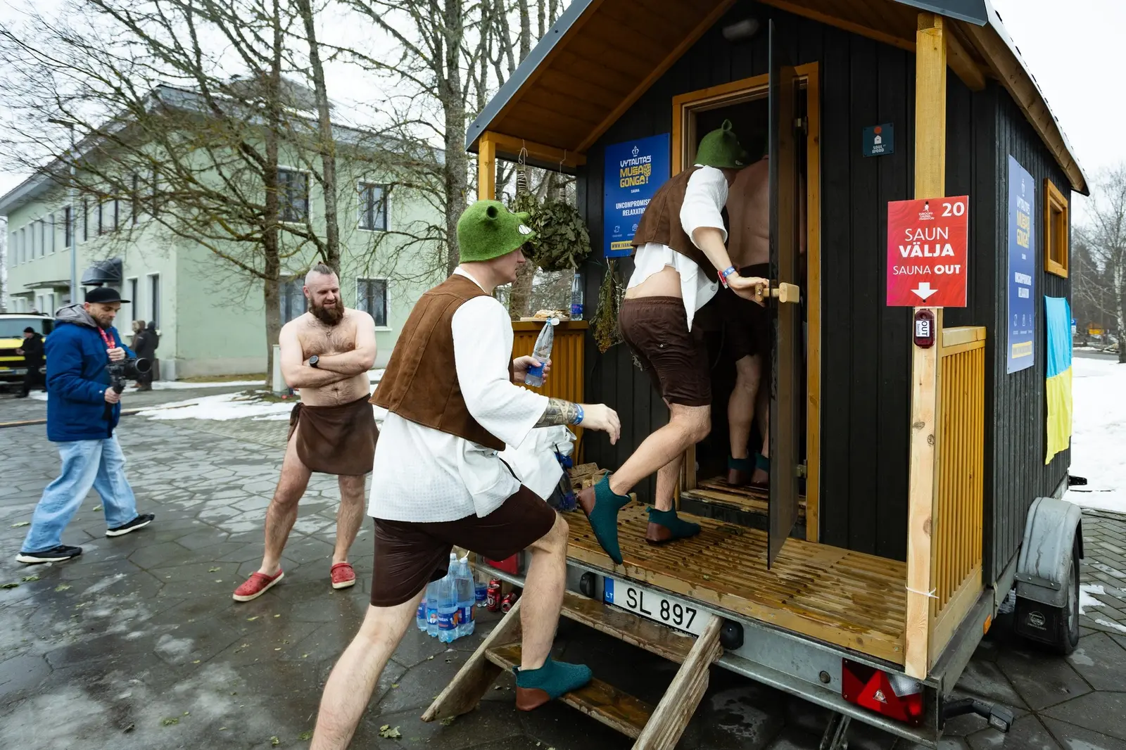 Costumed racers climbing the steps into a wooden trailer sauna parked on a snowy street in Otepää