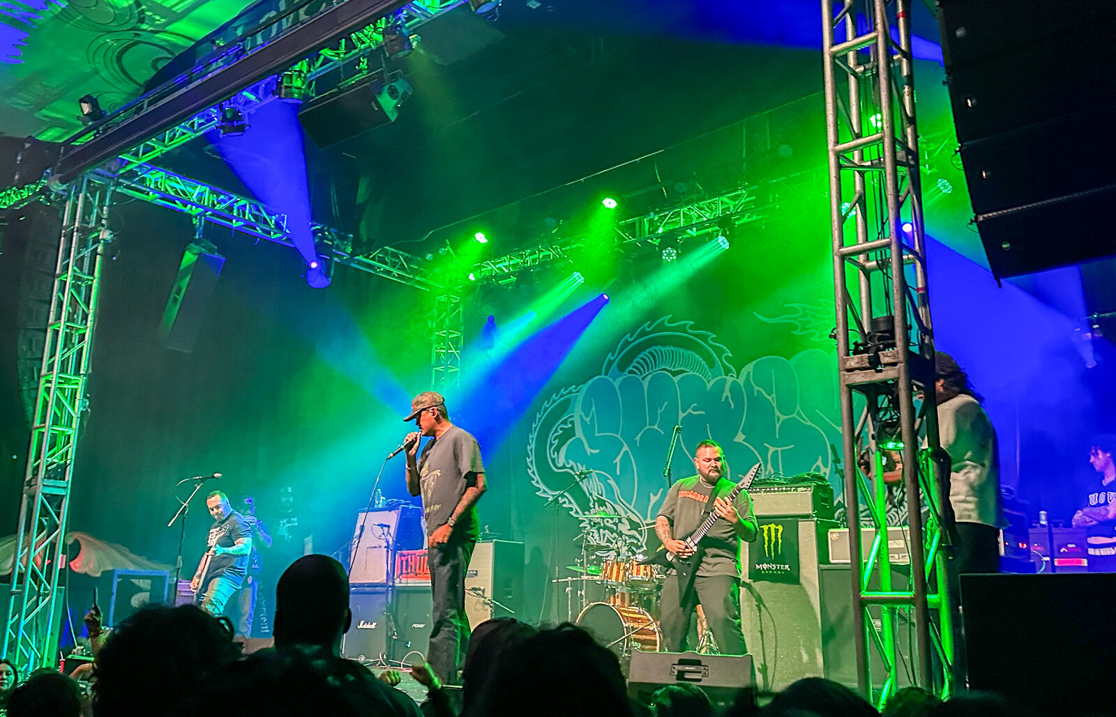 A band plays on a stage lit green and blue, with lighting trusses on both sides and a crowd in silhouette in the foreground