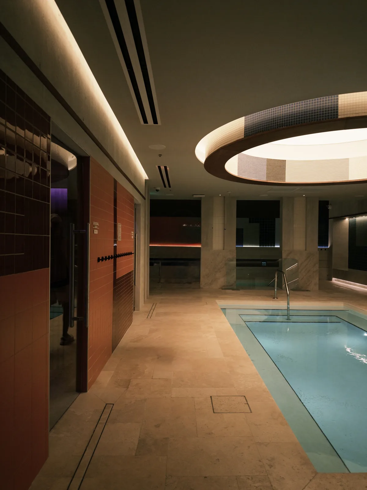 A long pool with a handrail beside a stone floor, under a round lit ceiling cove