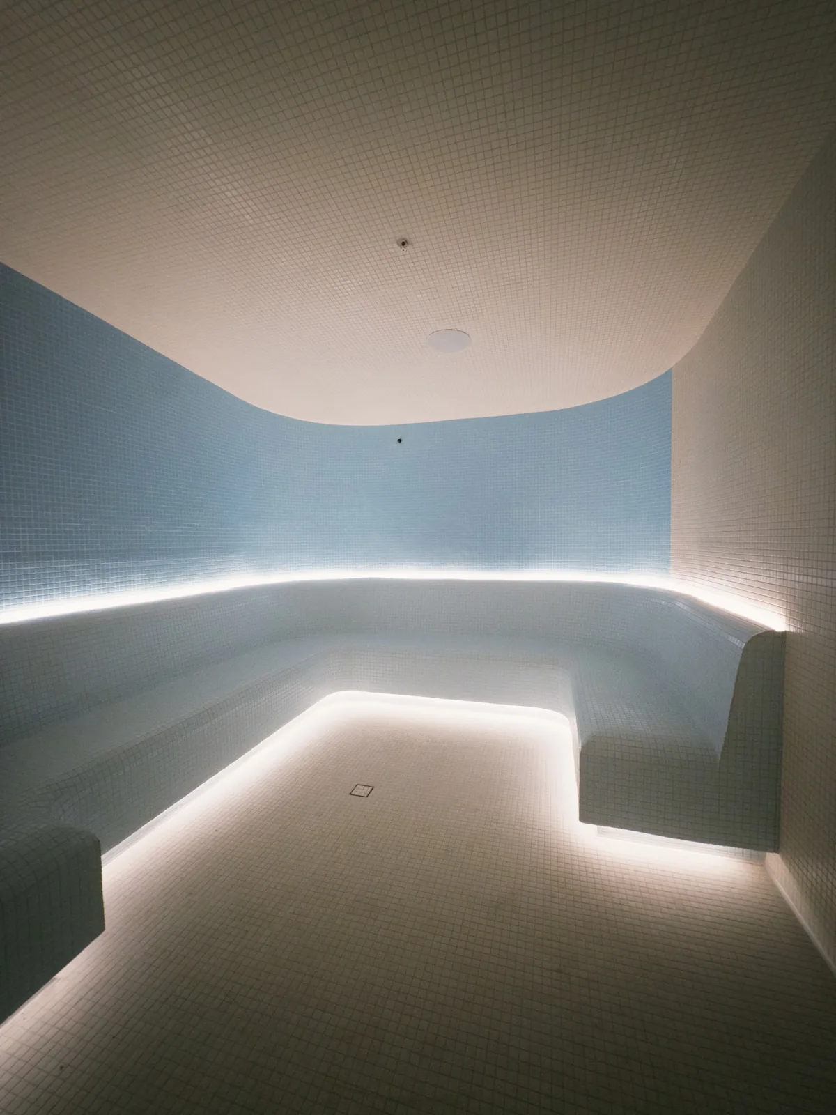 An empty steam room lined in pale blue and white mosaic tile, with a curved bench lit from below