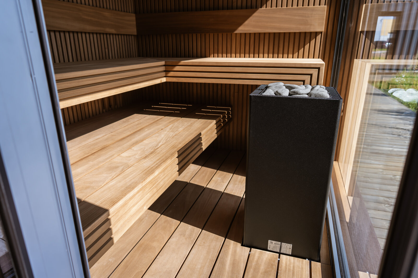 A tall black HUUM CORE Black heater filled with stones beside the wooden benches of a DWILD sauna with a glass front