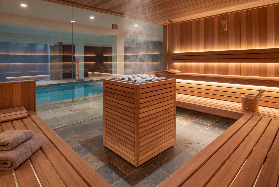 A HUUM CORE Pro heater clad in horizontal wooden slats with steam rising from its stones, in the middle of a lit wooden spa sauna with a pool behind a glass wall