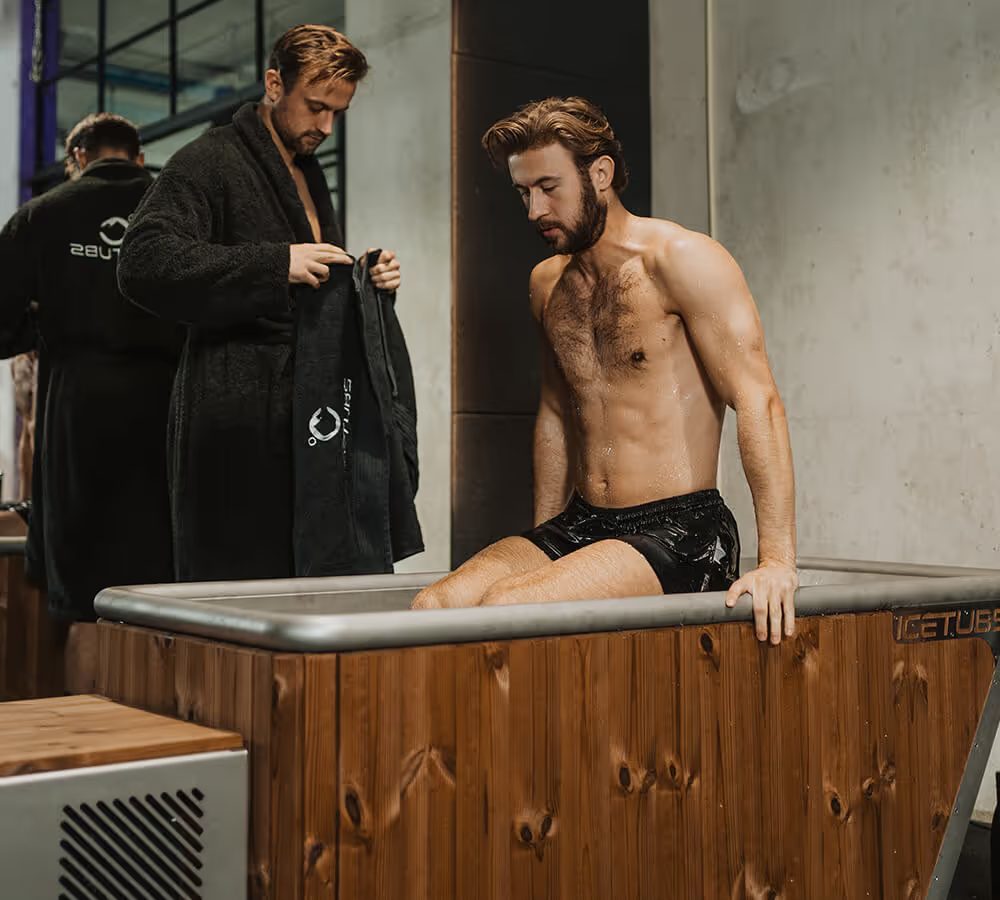 A man in swim shorts sitting on the rim of a wood-clad Icetubs cold tub while two people in robes stand behind him