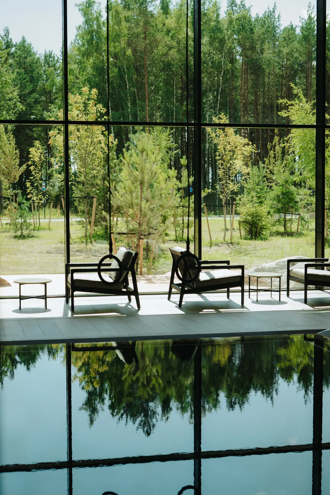 An indoor pool reflecting floor-to-ceiling windows, with lounge chairs on a terrace and young pine trees outside