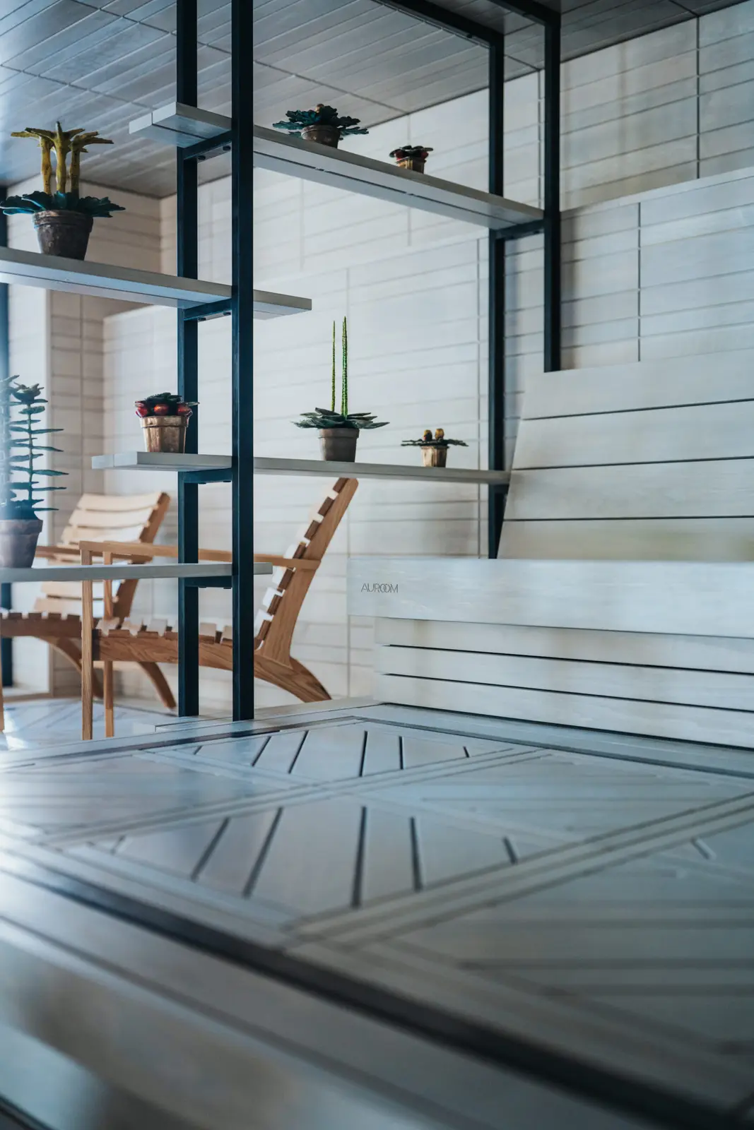 Wooden armchairs beside black metal shelves of potted plants in a pale wood-lined sauna with a diagonal slatted floor and white benches