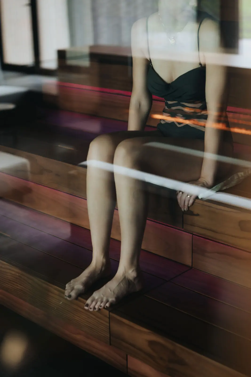 A bather in a black swimsuit sits on dark wooden sauna benches lit from below, seen through a glass wall