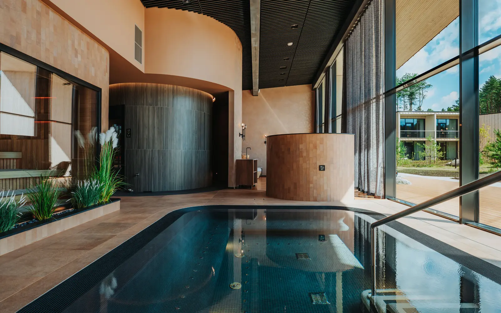 A narrow indoor pool beside tall windows, with a round wooden structure, plants and a curved wood-lined wall at the far end