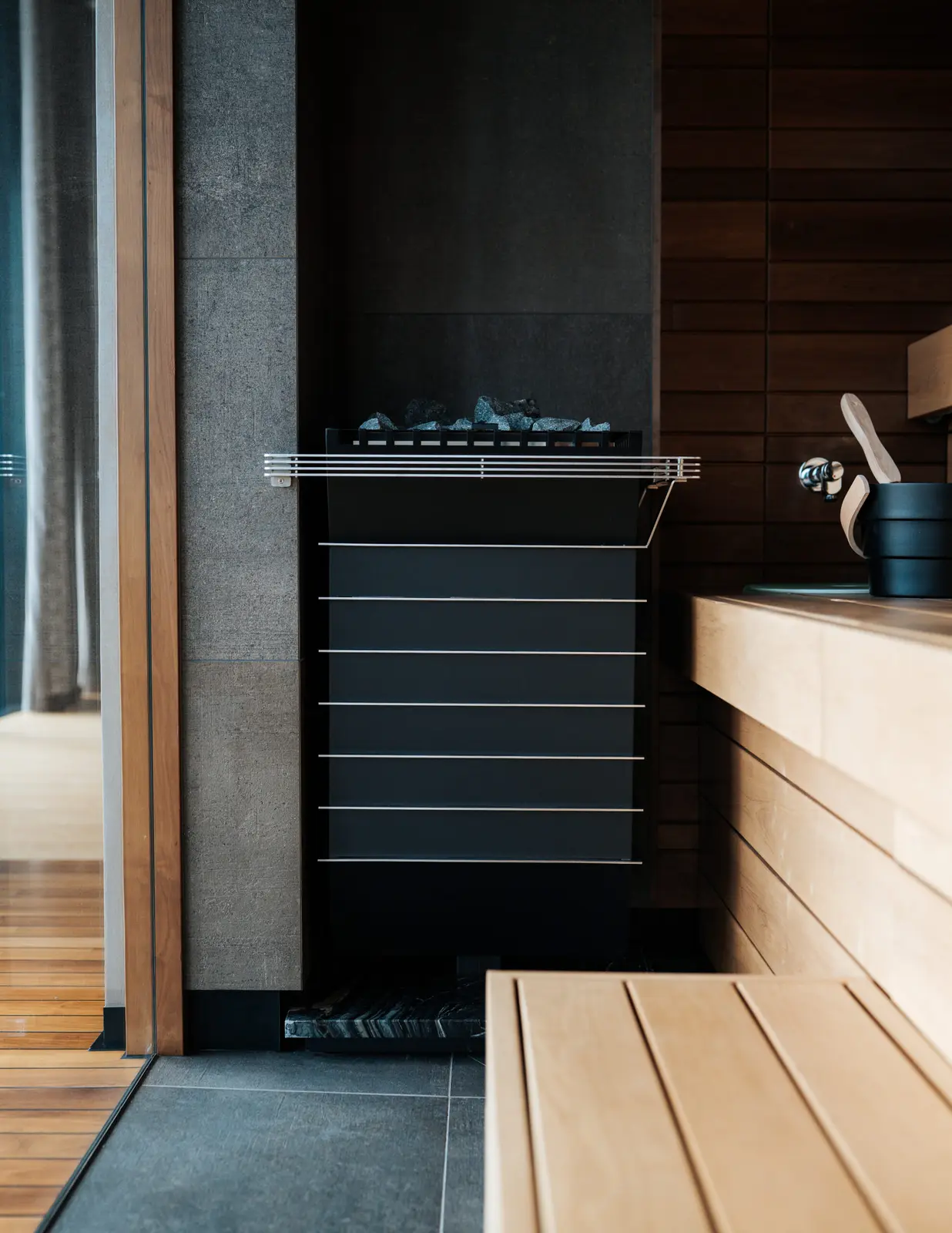 A black sauna heater topped with stones between a gray stone wall and pale wooden benches, with a wooden bucket and ladle on the bench