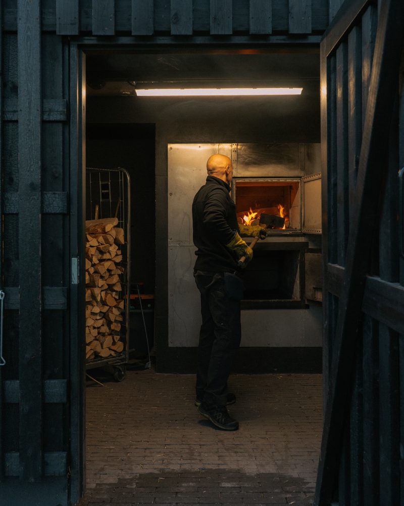 A stoker stands at a glowing firebox in a heating room stacked with firewood
