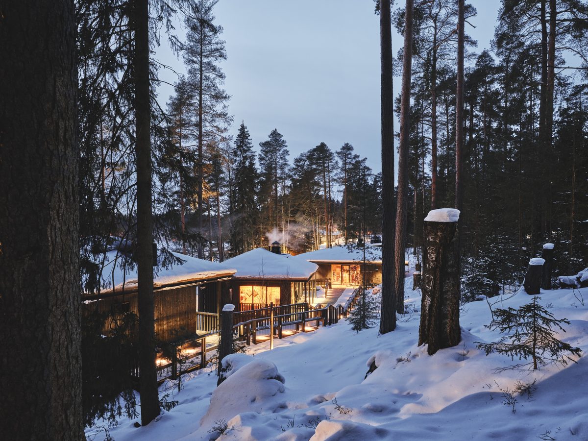 Lit timber sauna buildings among snowy pines at dusk