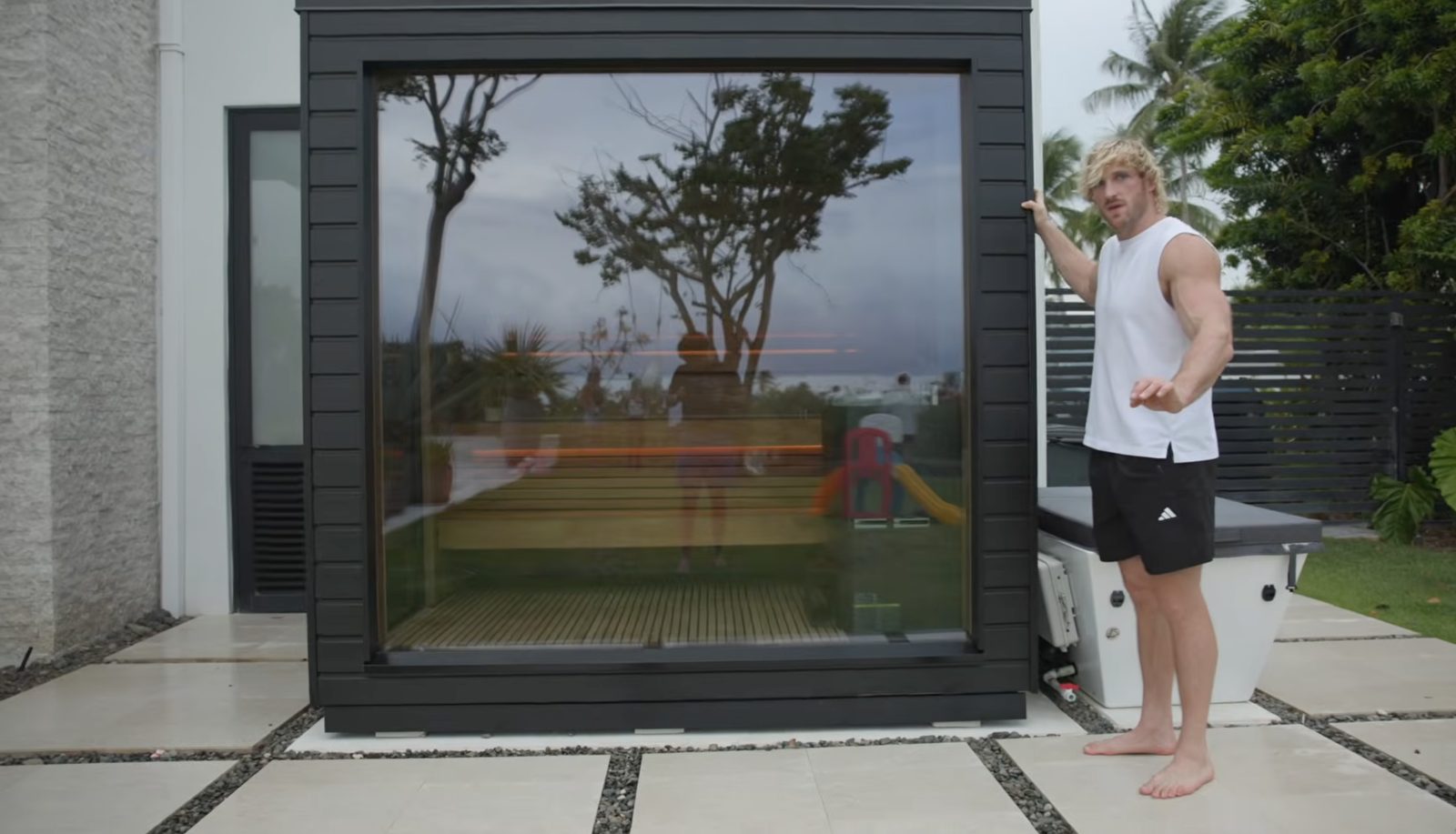 Logan Paul gesturing at his SaunaLife G6 outdoor sauna on his backyard patio with palm trees in background