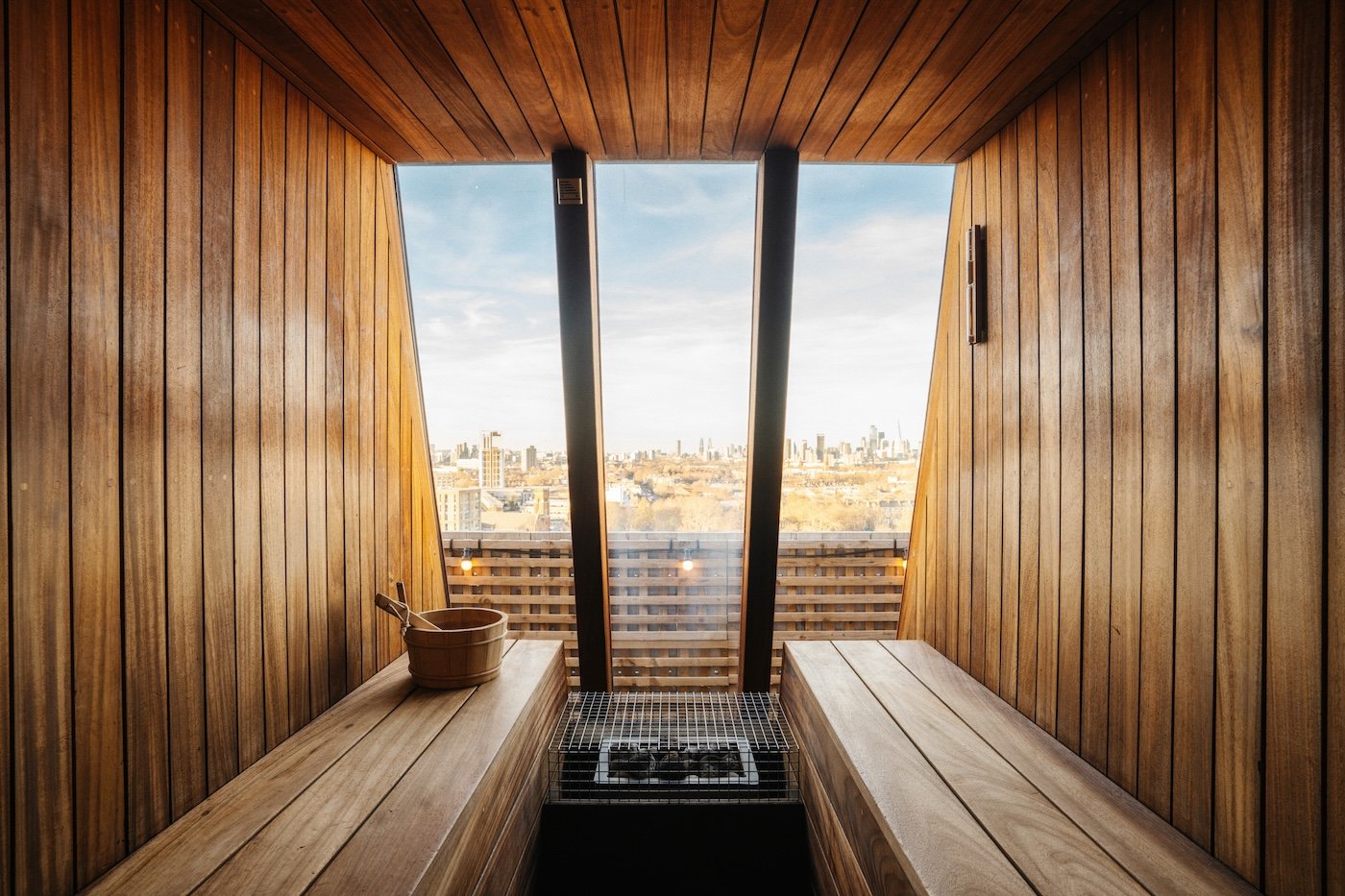 A wood-lined sauna with benches on either side of a stove and a tall window onto a city skyline
