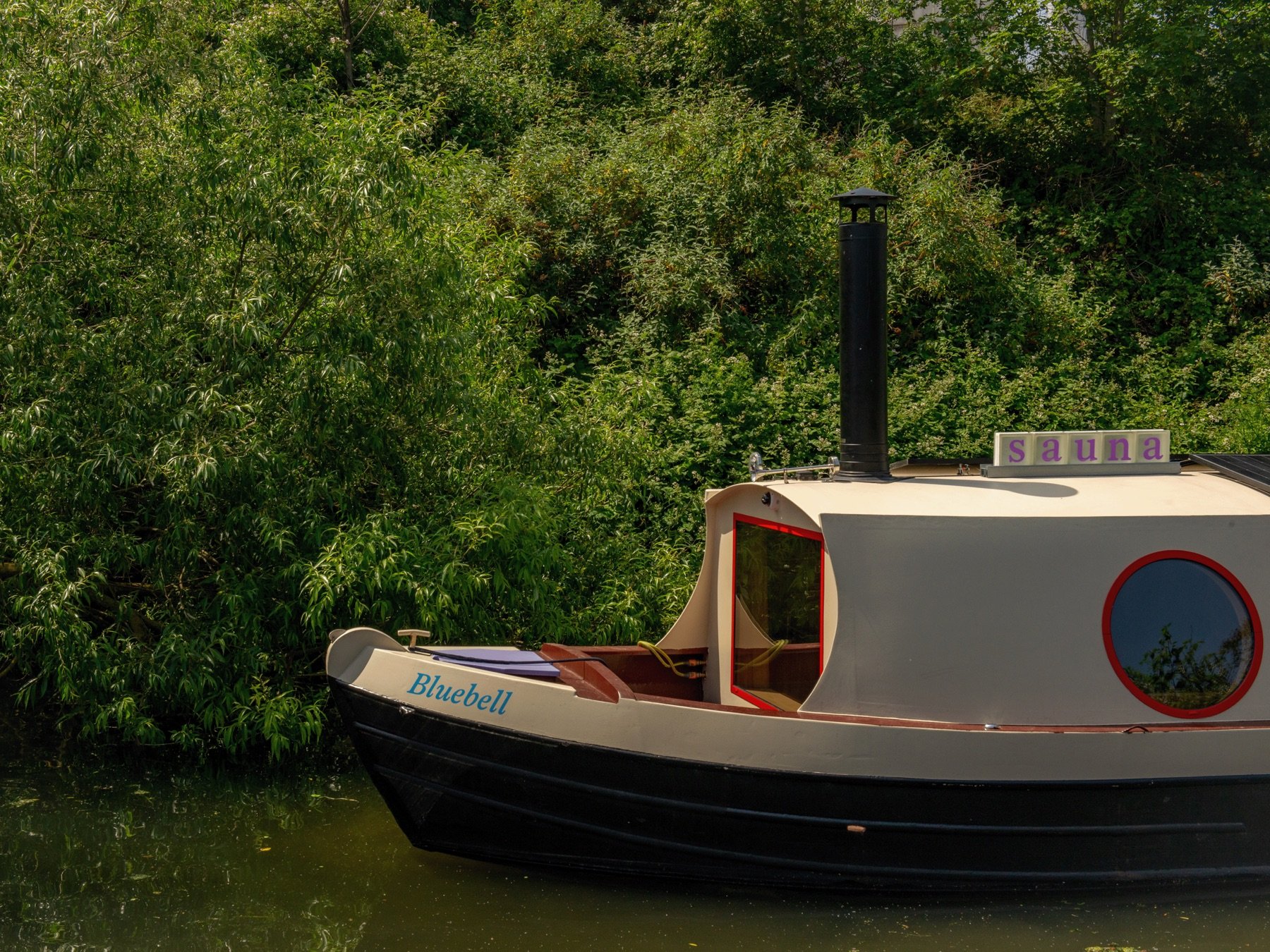A small boat named Bluebell, with a stovepipe chimney and a sauna sign on its roof, against a bank of green trees