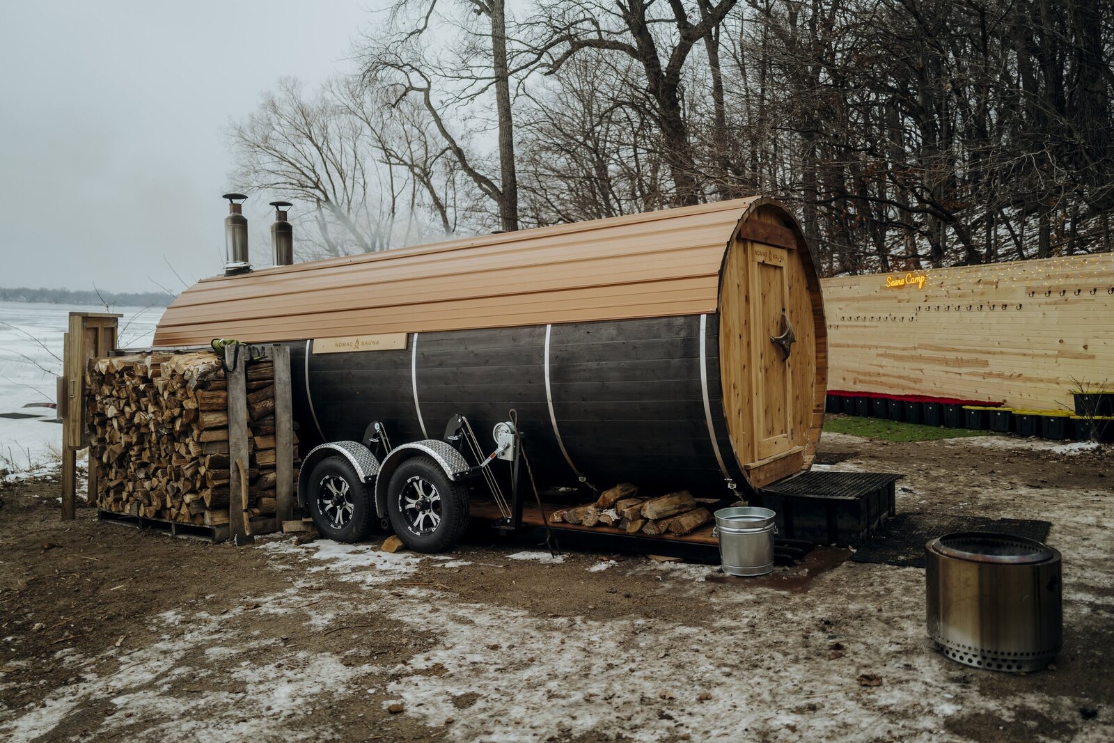 A long barrel sauna with two stovepipes on a dual-axle trailer beside a frozen lake, with split firewood stacked at the hitch end and a metal ash bucket by the door