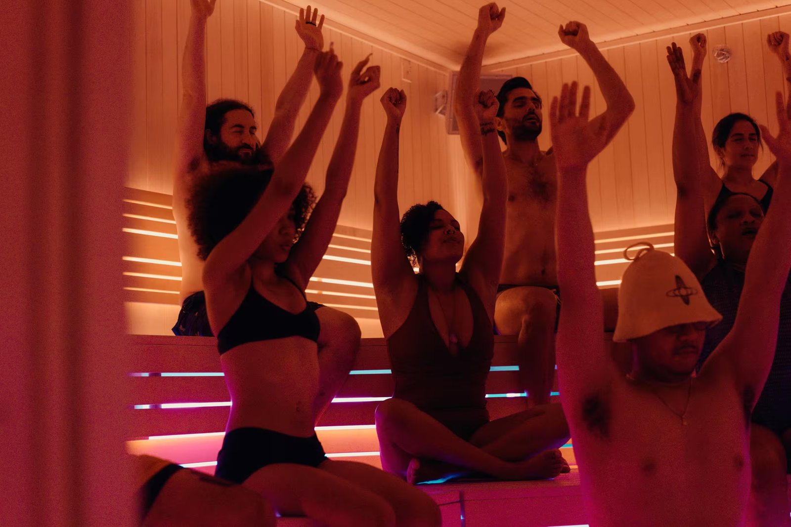 Guests on tiered sauna benches raise their arms with eyes closed during a guided session in warm orange light