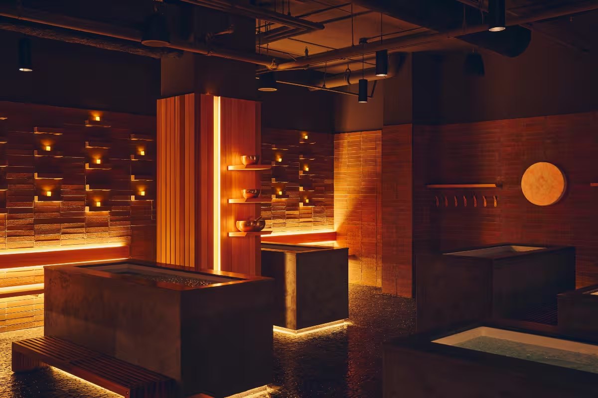 Several concrete cold plunge tubs in a dim, warm-lit room with brick walls, wood columns and candles in wall niches