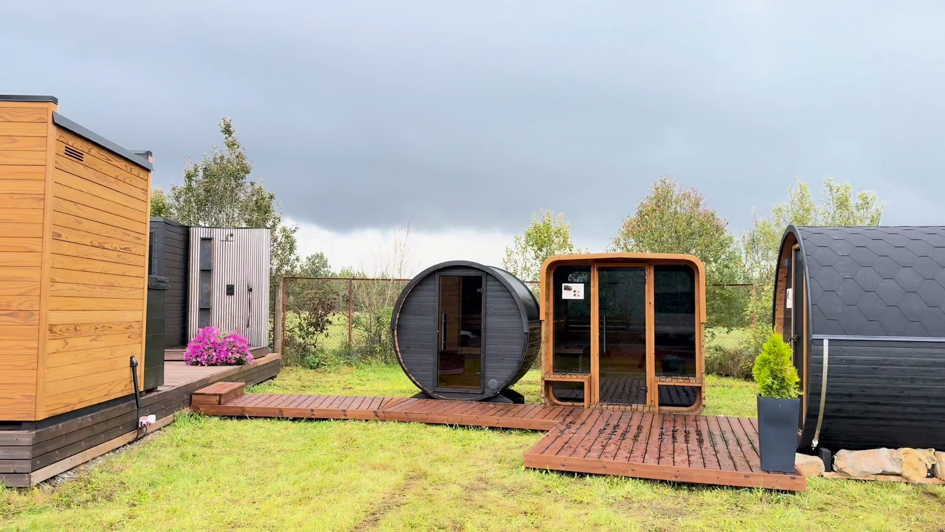 Barrel saunas, a timber cabin and a glass-fronted sauna on a deck in a factory yard under a heavy gray sky
