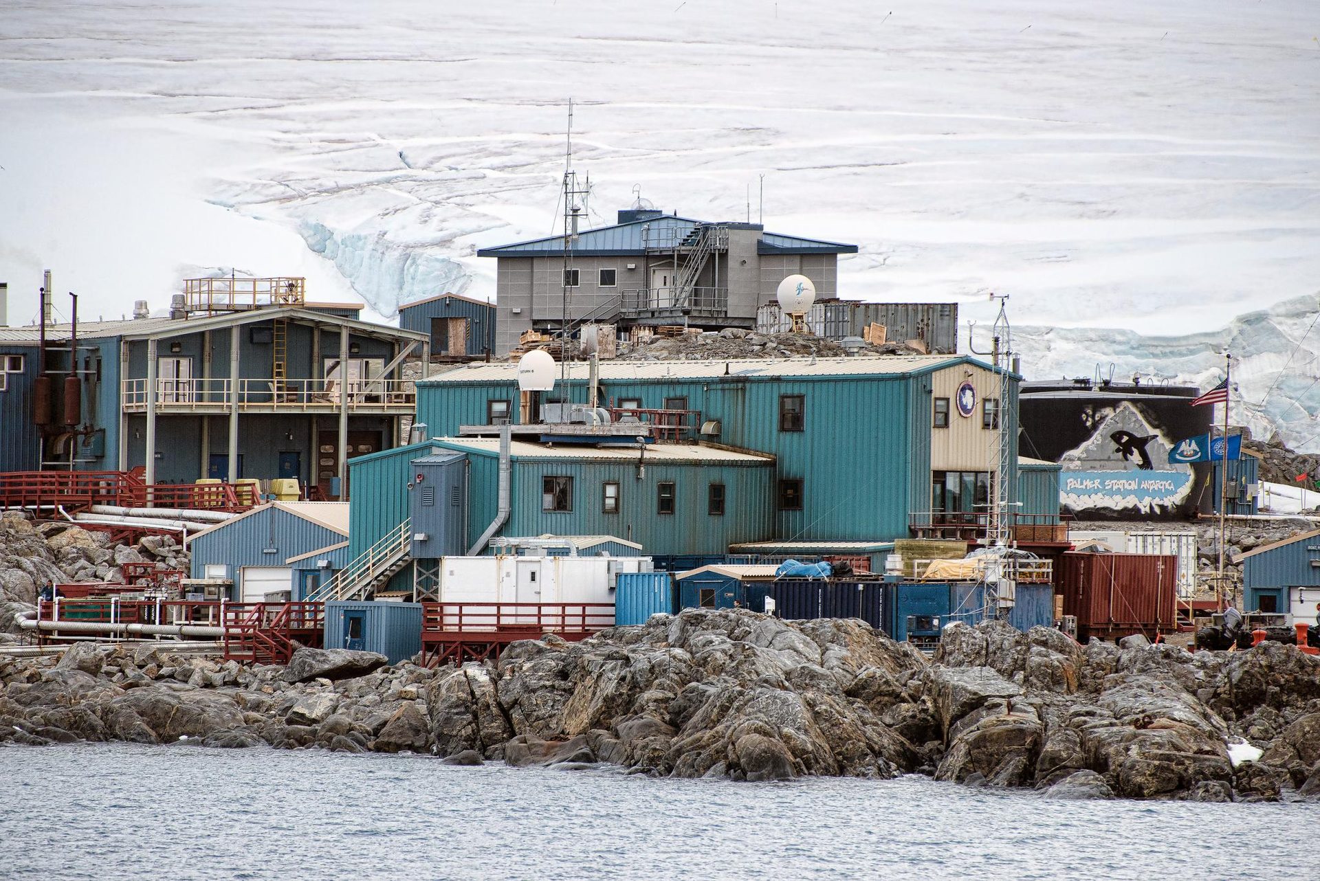 Palmer Station, Antarctica, a cluster of blue and tan buildings on a rocky shore with a glacier behind